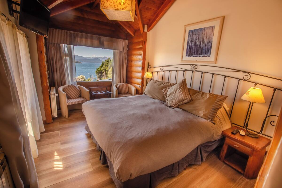 Un dormitorio con una cama grande, dos mesitas de noche con lámparas, una silla, vistas al mar y un techo de madera.
