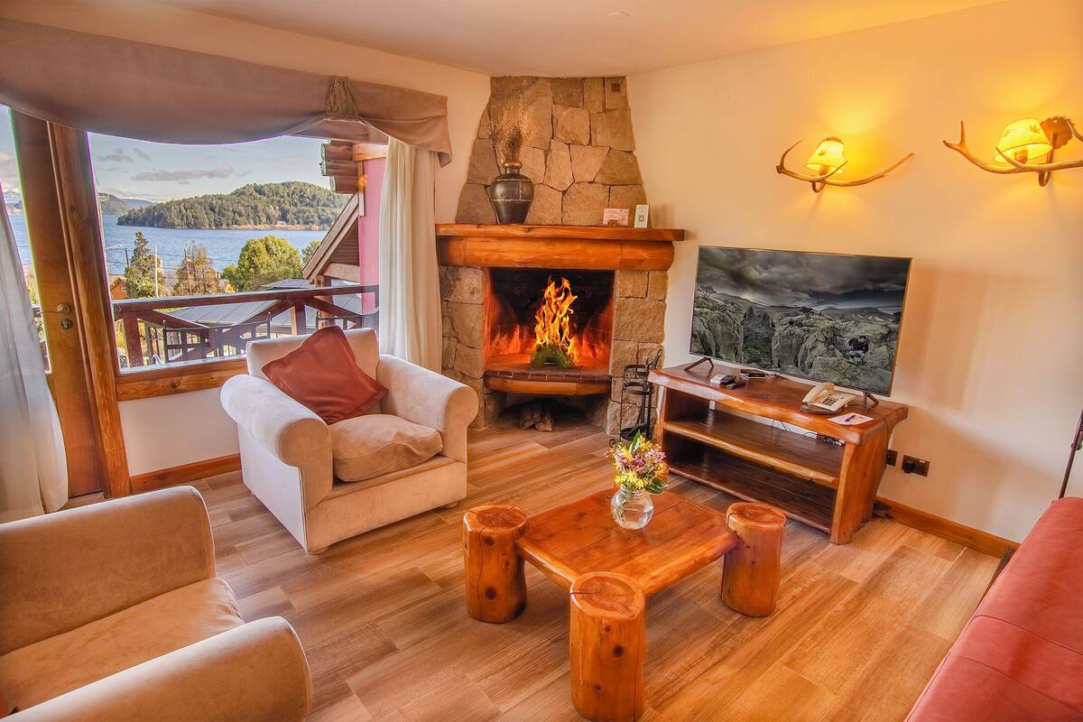Una sala de estar acogedora con chimenea de piedra, muebles de madera, un televisor y una vista panorámica de un lago y montañas.