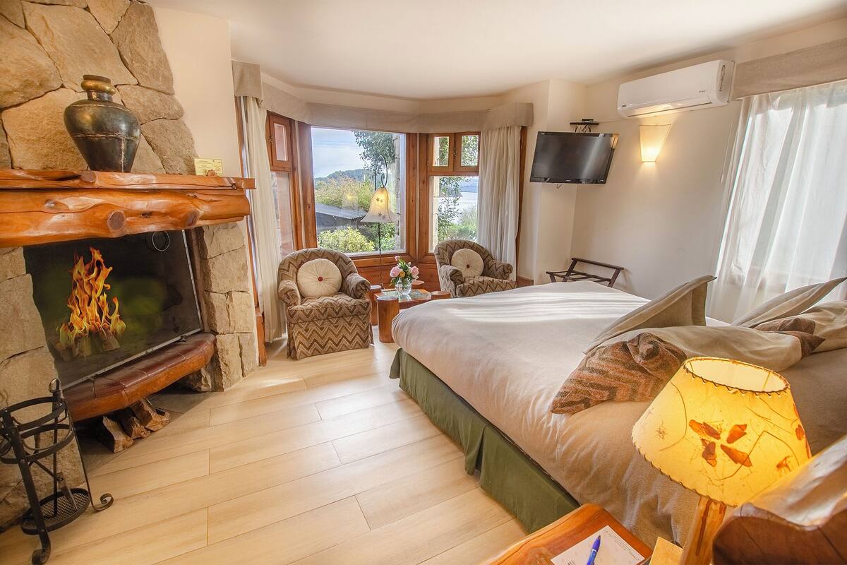 Un dormitorio acogedor con chimenea de piedra, una cama con colcha verde, dos sillones, una mesita de noche de madera y un televisor montado en la pared.