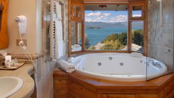 Un baño con una bañera de madera, un ventanal con vistas a un lago y montañas, y un secador de pelo en la pared.