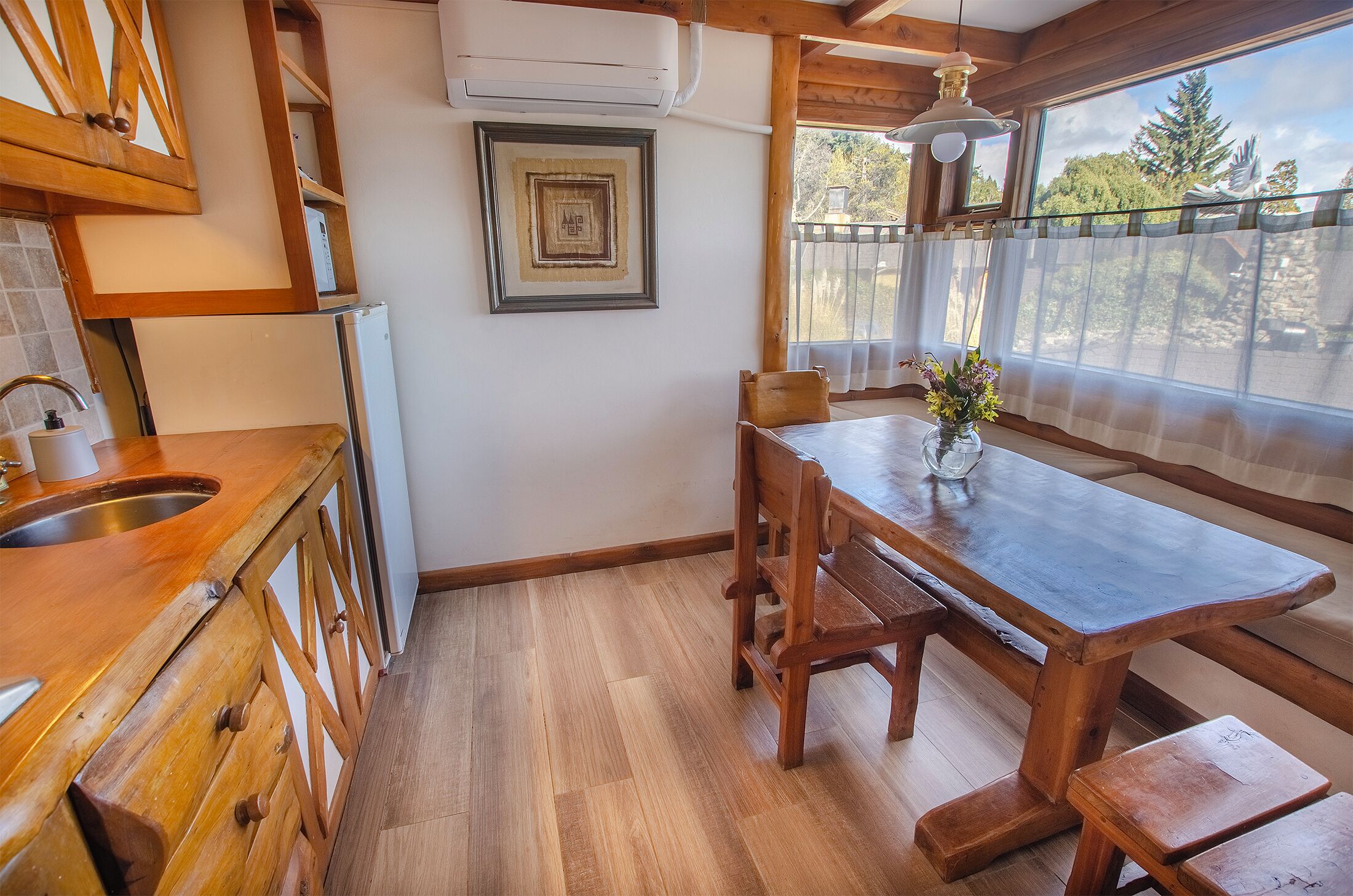 A kitchen with wooden cabinets, a built-in refrigerator, a wooden dining table with benches, and a window with a view of trees.