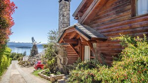 Una cabaña de madera con chimenea de piedra, vista a un lago y un coche rojo estacionado al frente.