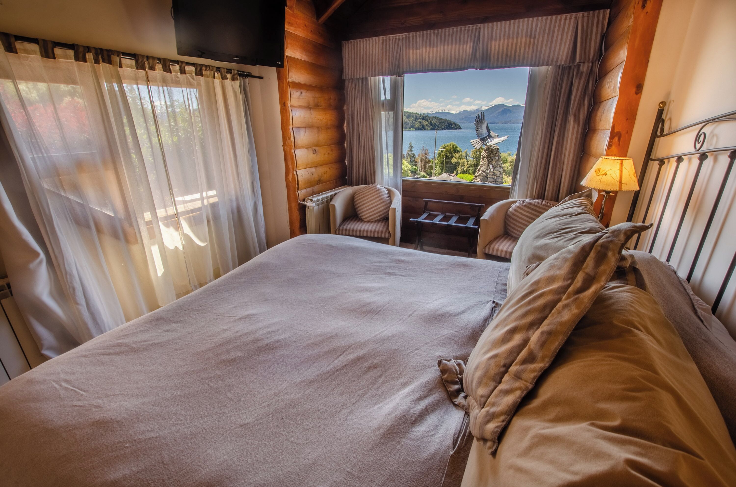 A log cabin room with a large bed, a chair, a small table, and a view of a lake and mountains.