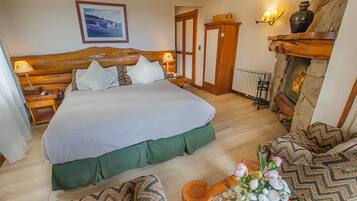 A bedroom with a large bed, wooden headboard, bedside lamps, a wooden wardrobe, a stone fireplace, and a framed picture on the wall.