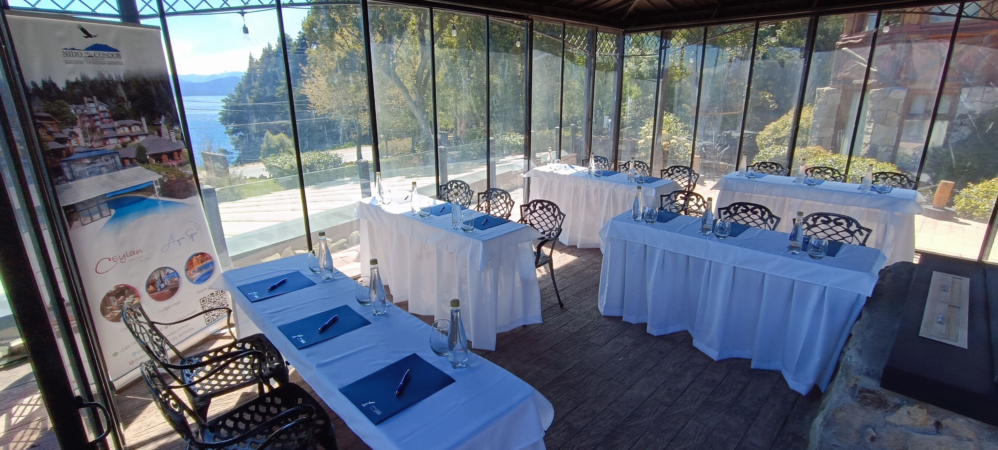 A restaurant with outdoor seating, white tablecloths, and black metal chairs. Tables are set with blue napkins, water bottles, and glasses. A large poster with a scenic view is visible on the left.