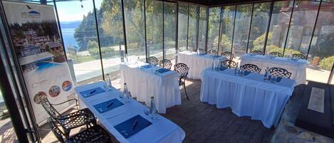 Een restaurant met terras, witte tafellakens en zwarte metalen stoelen. De tafels zijn gedekt met blauwe servetten, waterflessen en glazen. Een grote poster met een mooi landschap is links te zien.