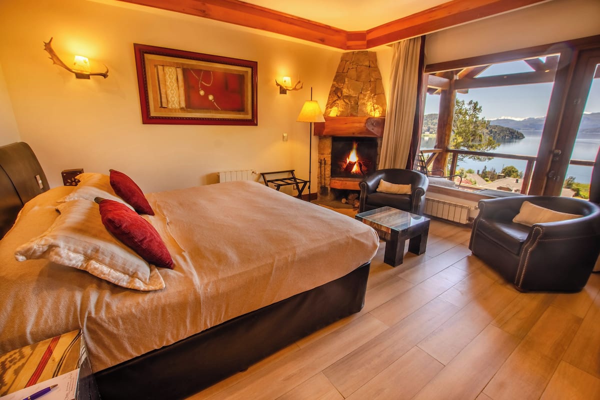 Habitación de hotel con una cama grande, chimenea, vista al lago y un rincón de descanso acogedor.
