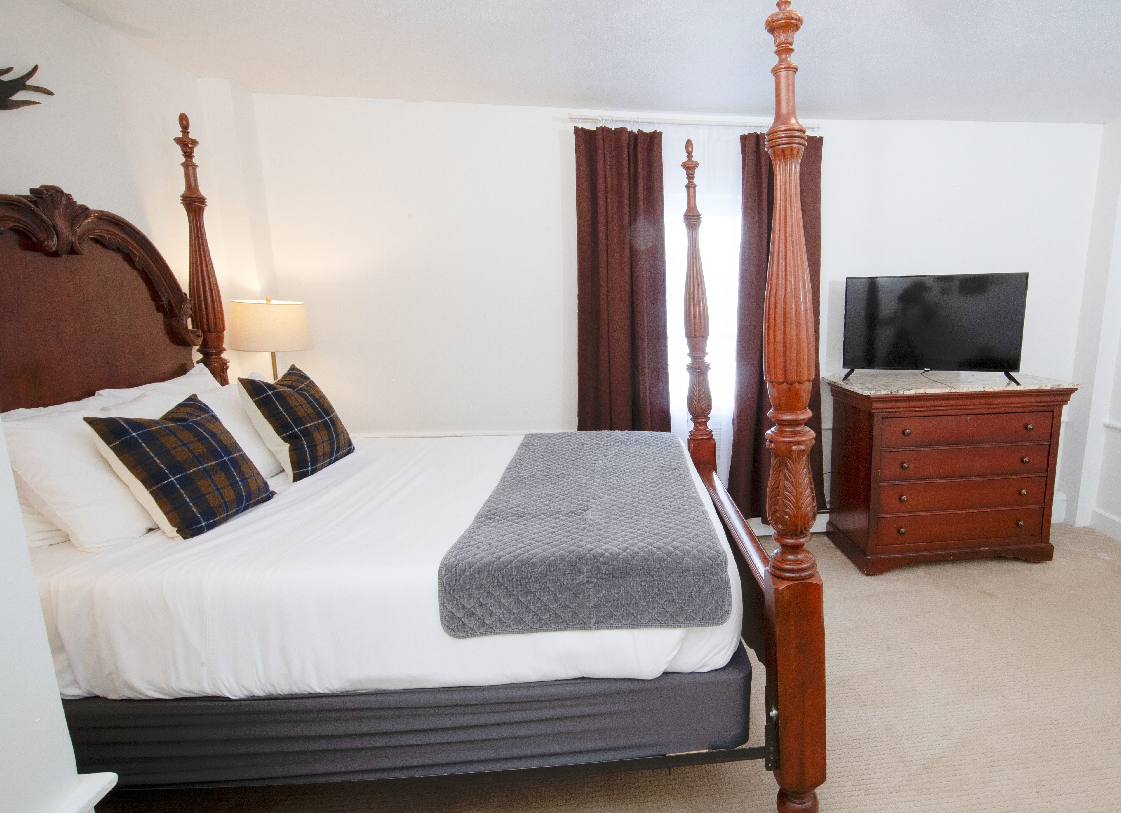 A four-poster bed with a wooden headboard, a television on a wooden dresser, and a lamp on a bedside table.