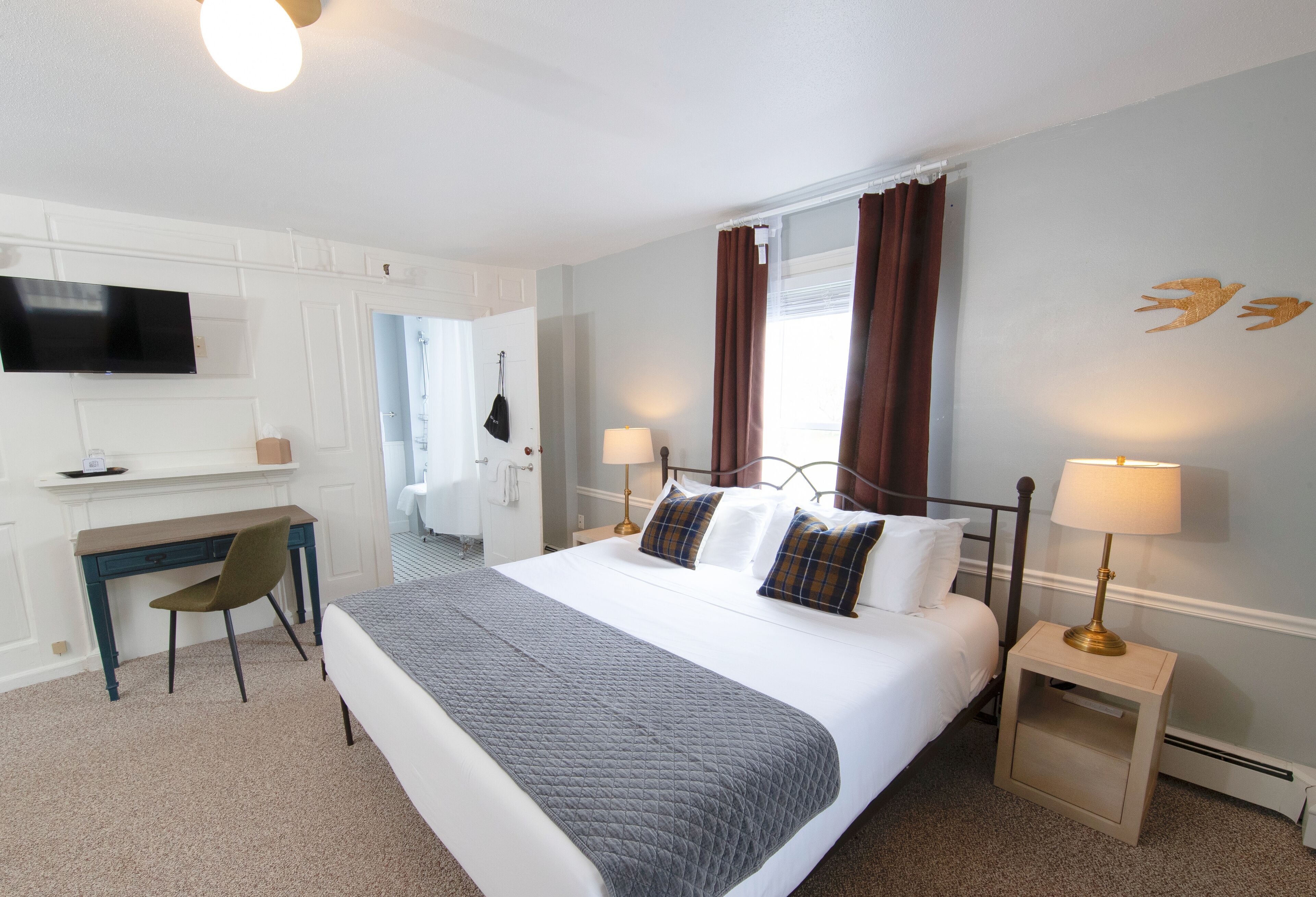Habitación de hotel con cama, escritorio, silla, televisor, lámparas y cortinas.