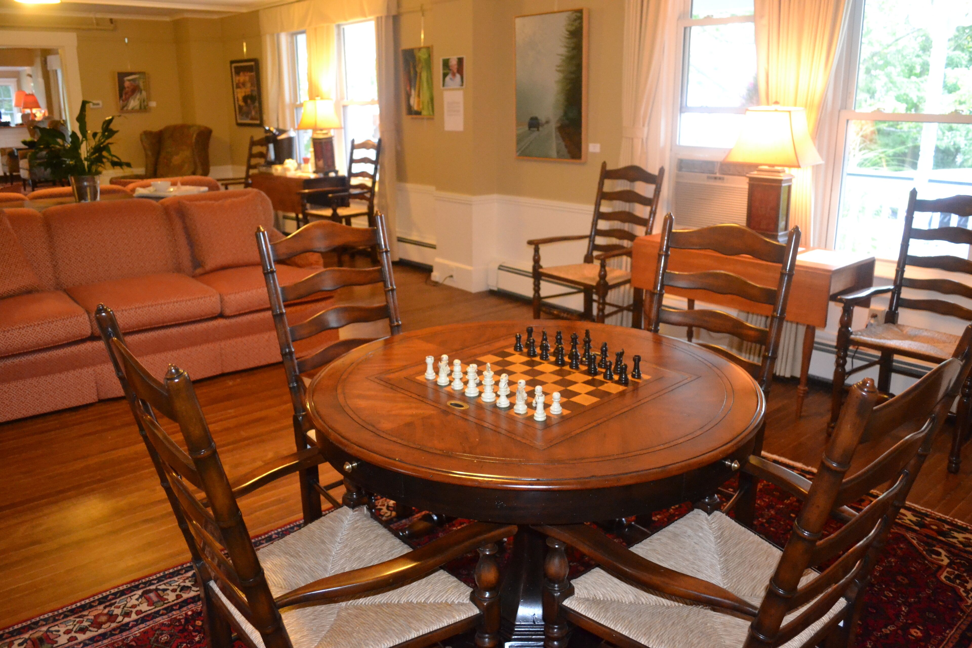 A room with a round wooden table set for a game of chess, surrounded by wooden chairs with cushioned seats.