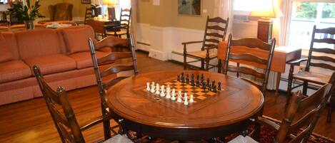 A room with a round wooden table set for a game of chess, surrounded by wooden chairs with cushioned seats.