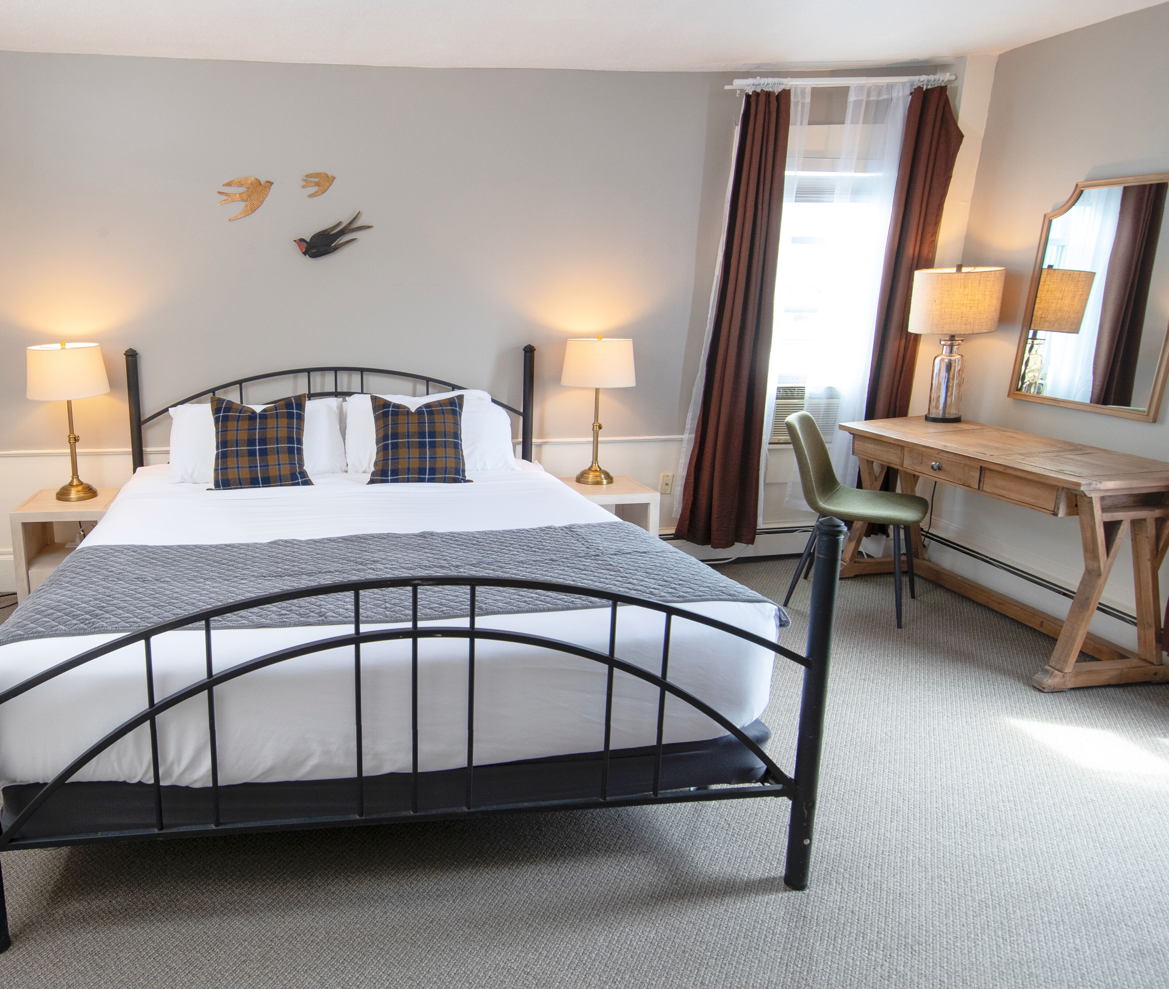 A hotel room with a bed, two bedside tables with lamps, a desk with a chair, a mirror, and a window with curtains.