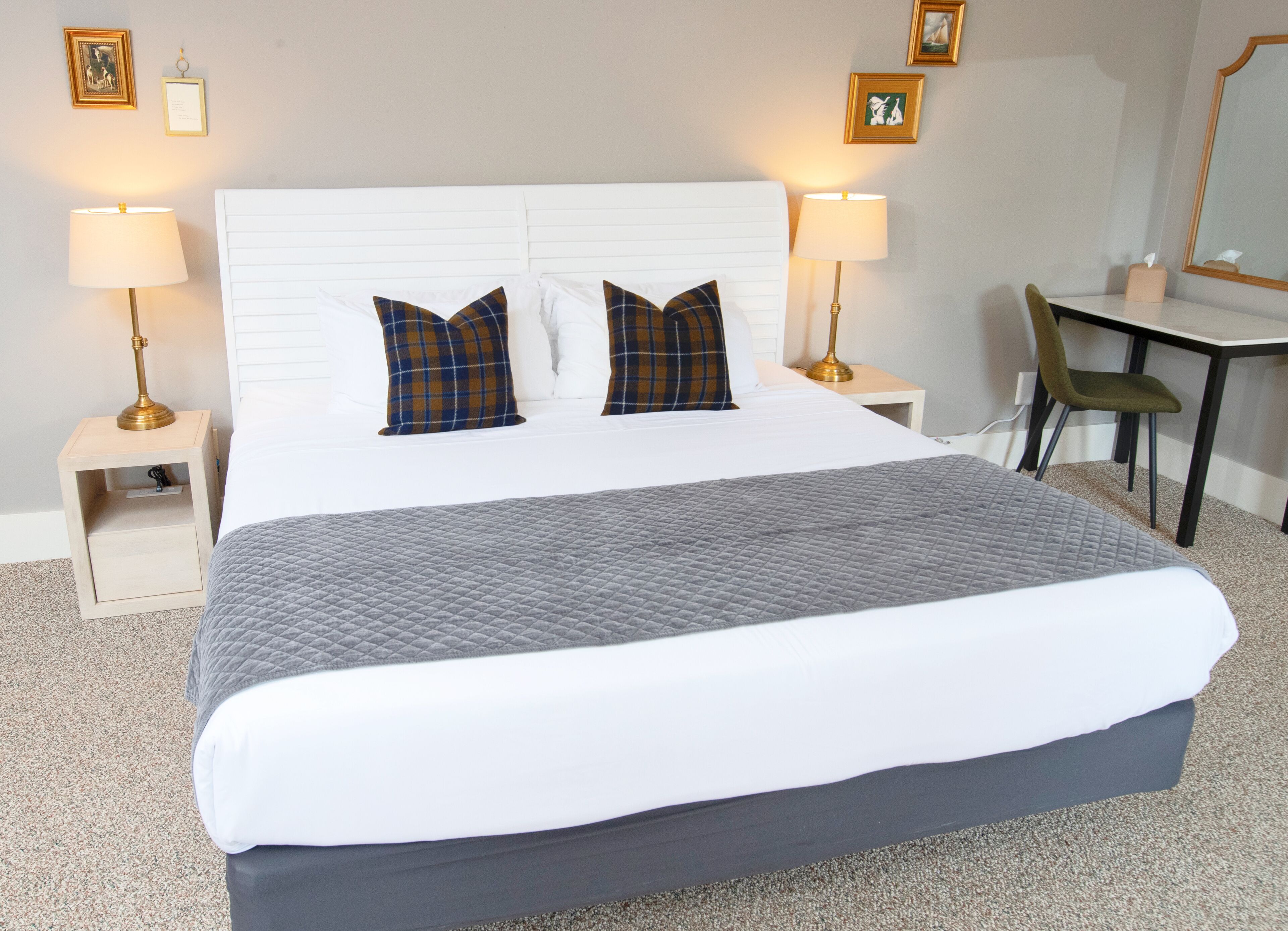 Een net opgemaakt bed met een grijze gewatteerde deken, twee geruite kussens en een wit hoofdbord. Er staan twee bedlampen met gouden voetstukken, een klein nachtkastje met een lade, en een groene stoel naast een bureau met een spiegel en een ingelijst beeld aan de muur.