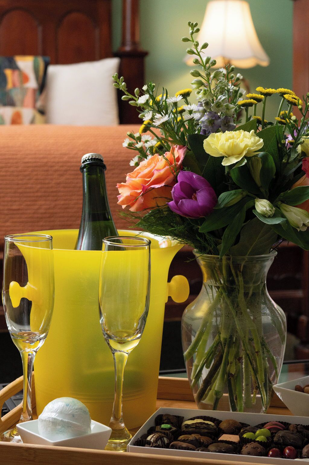 A tray with a vase of flowers, two glasses of a yellow beverage, a bottle, and a box of chocolates.