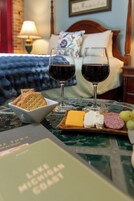 A hotel room with a bed, a bedside table with a lamp, a tray with cheese, crackers, and grapes, and two glasses of red wine.