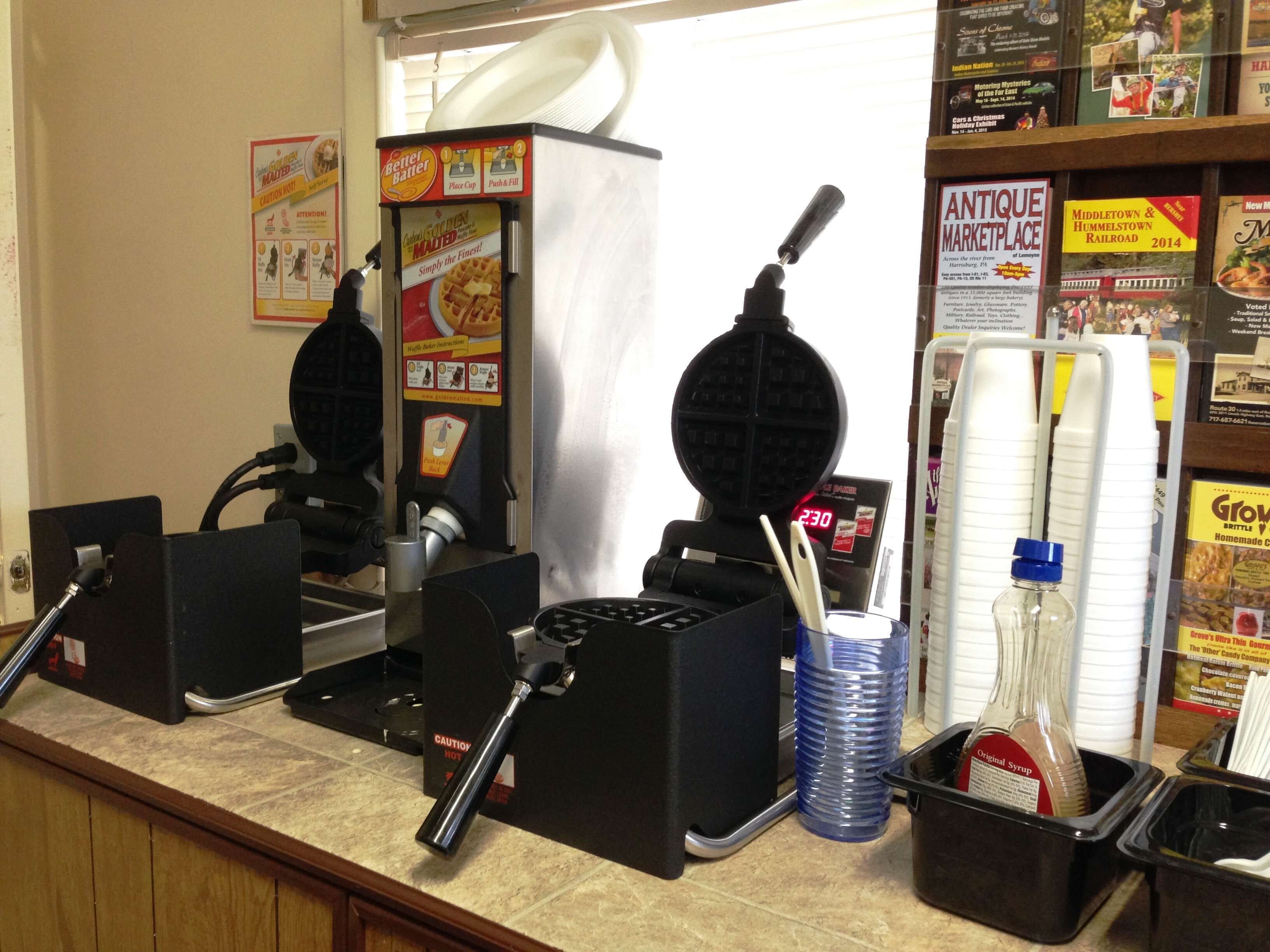 A waffle maker, a bottle of syrup, and a menu board in a restaurant setting.