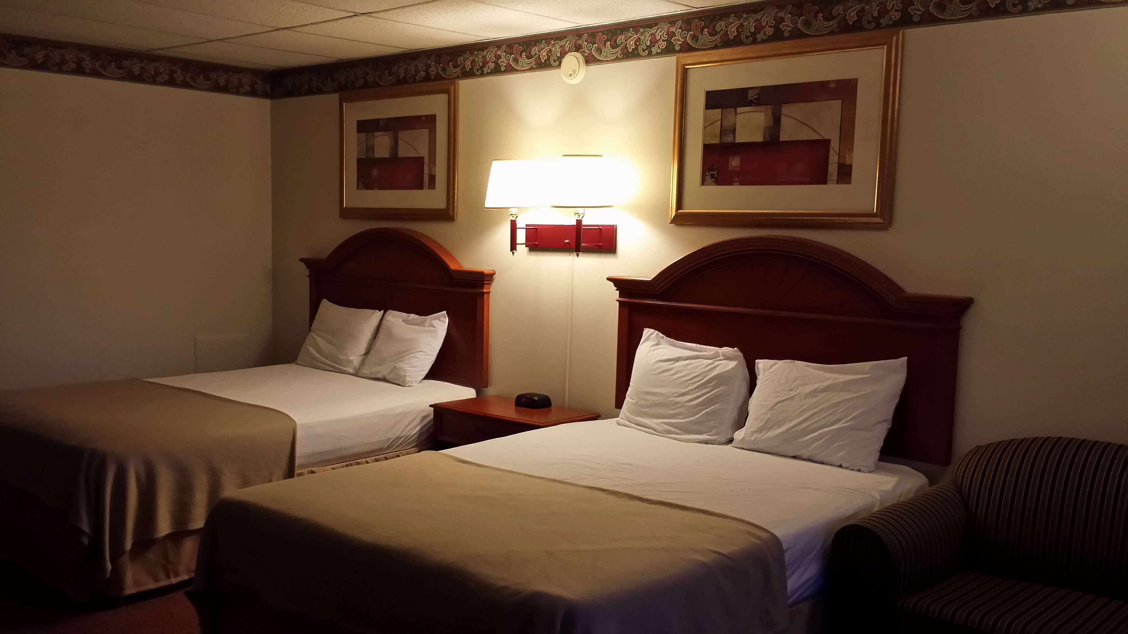 A hotel room with two single beds, a nightstand with a lamp, a chair, and two framed pictures on the wall.