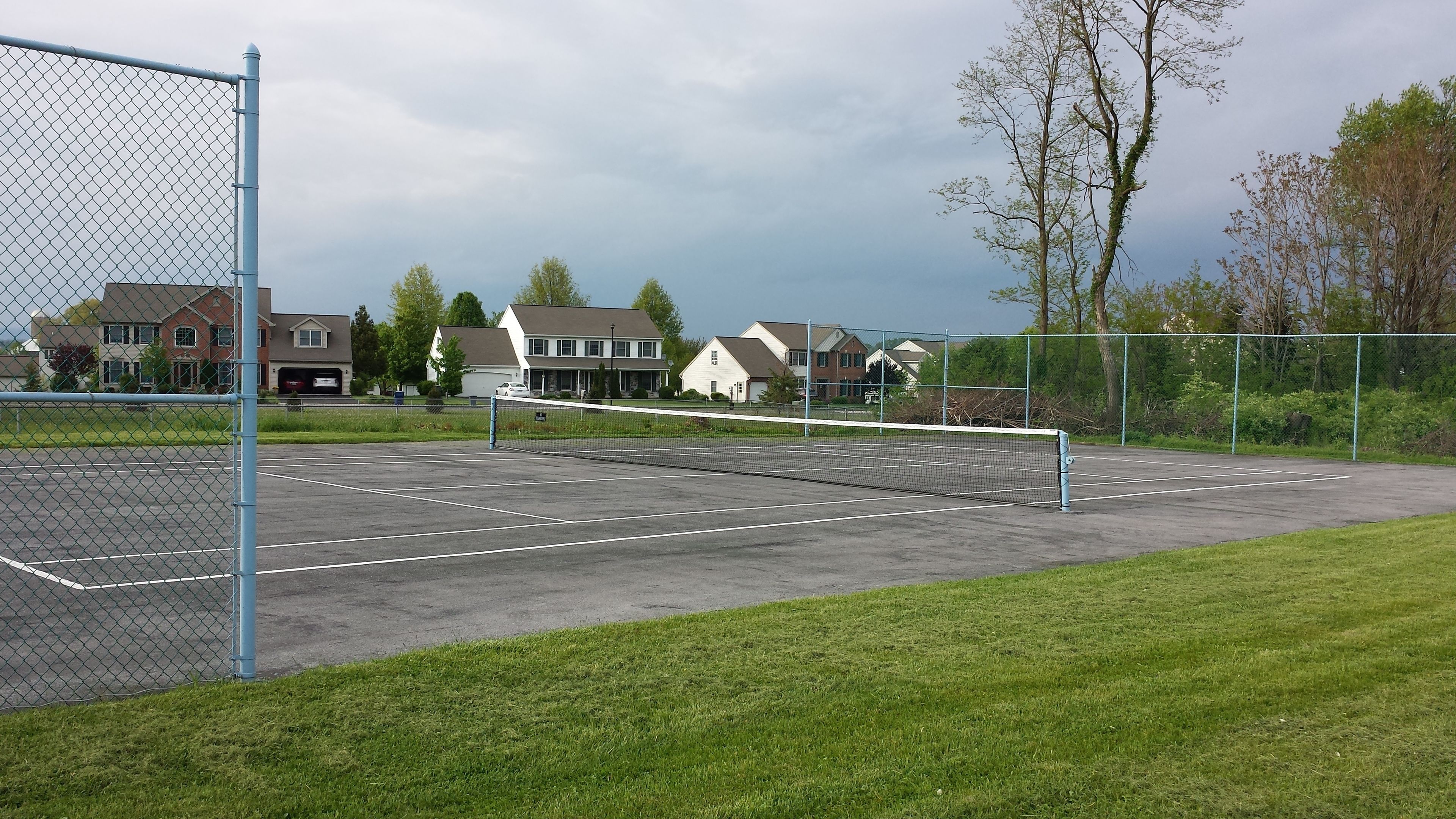 A tennis court with a net, surrounded by a chain-link fence, and houses in the background.