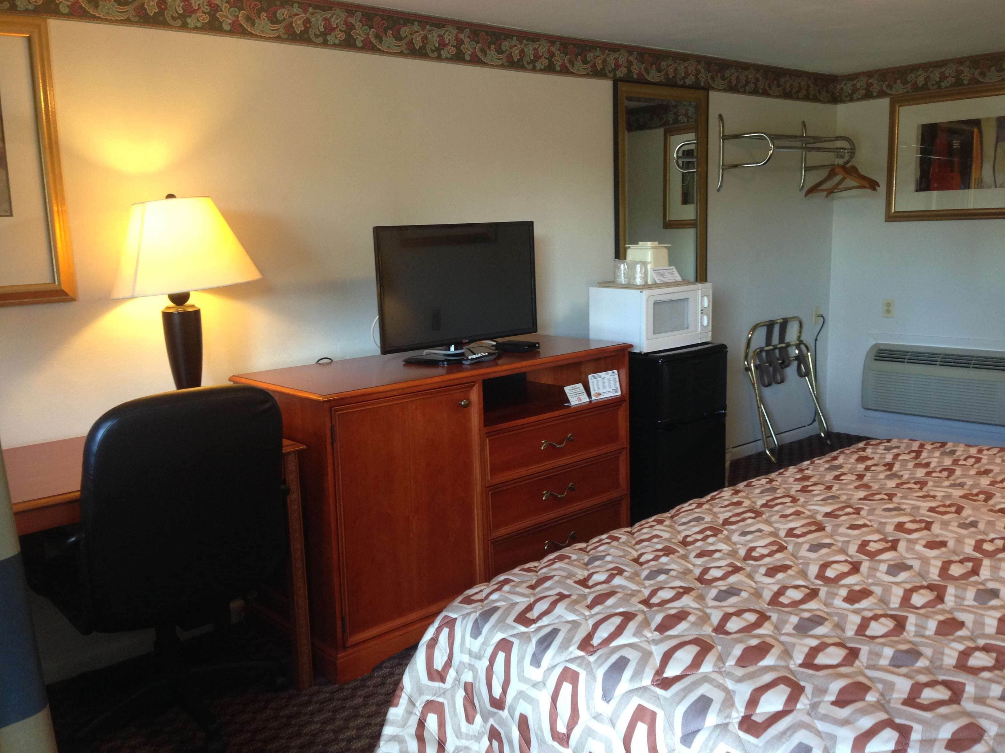 Habitación de hotel con escritorio de madera, televisor, microondas, refrigerador, cama con cubrecama estampado, silla, lámpara, espejo y perchero.