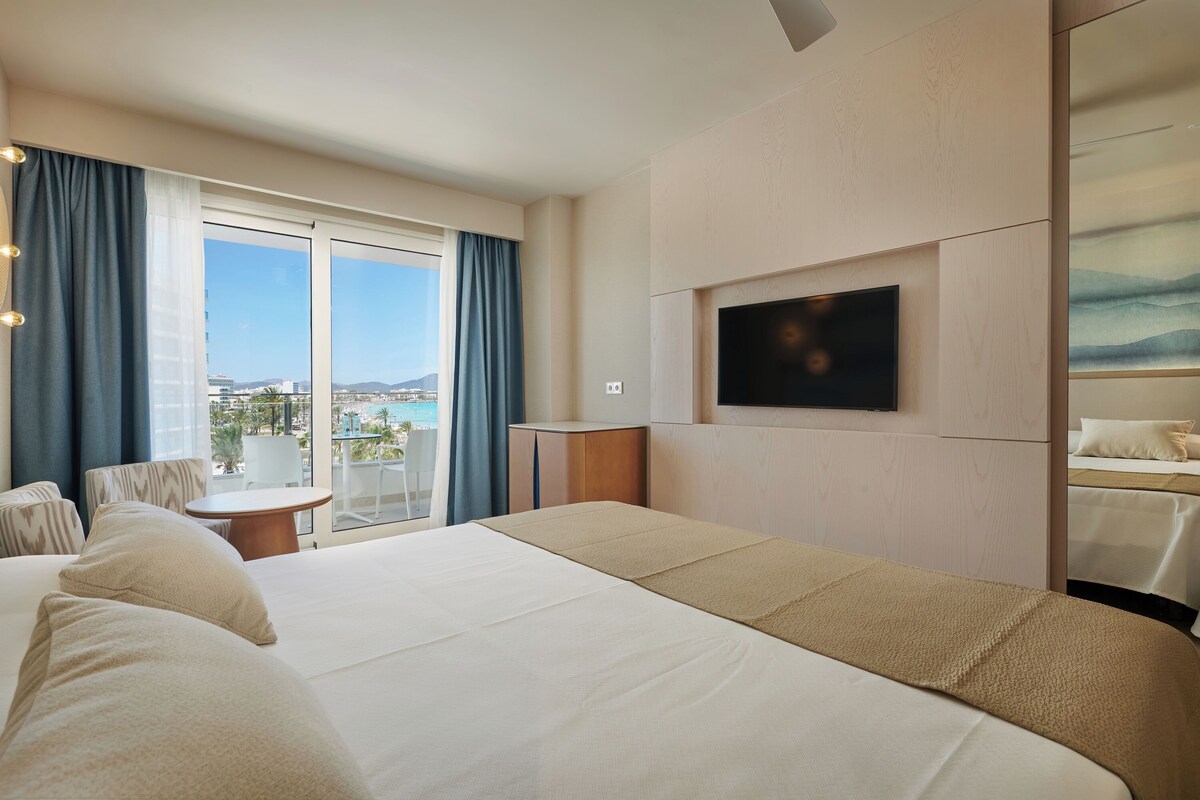Une chambre d’hôtel dotée d’un grand lit, d’un téléviseur fixé au mur, d’un balcon offrant une vue sur la ville et d’un coin salon avec une table et des chaises.