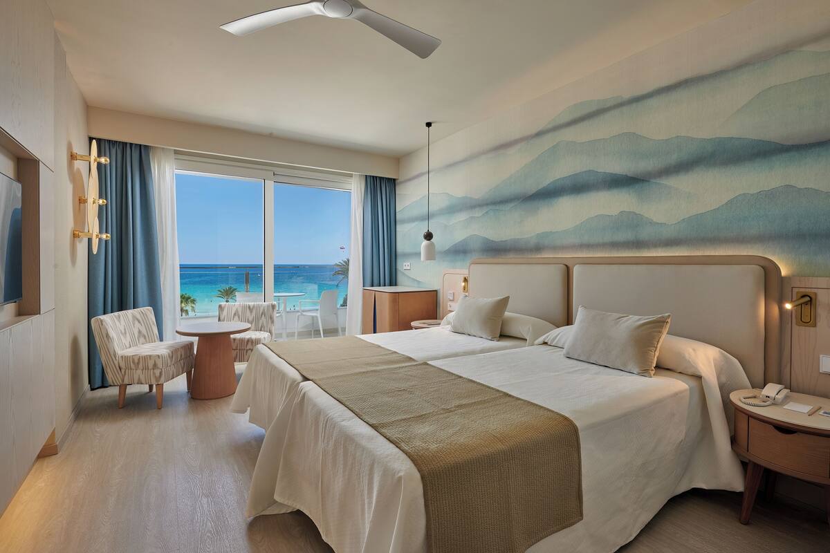 A hotel room with two beds, a view of the ocean, a ceiling fan, and a painting of mountains.