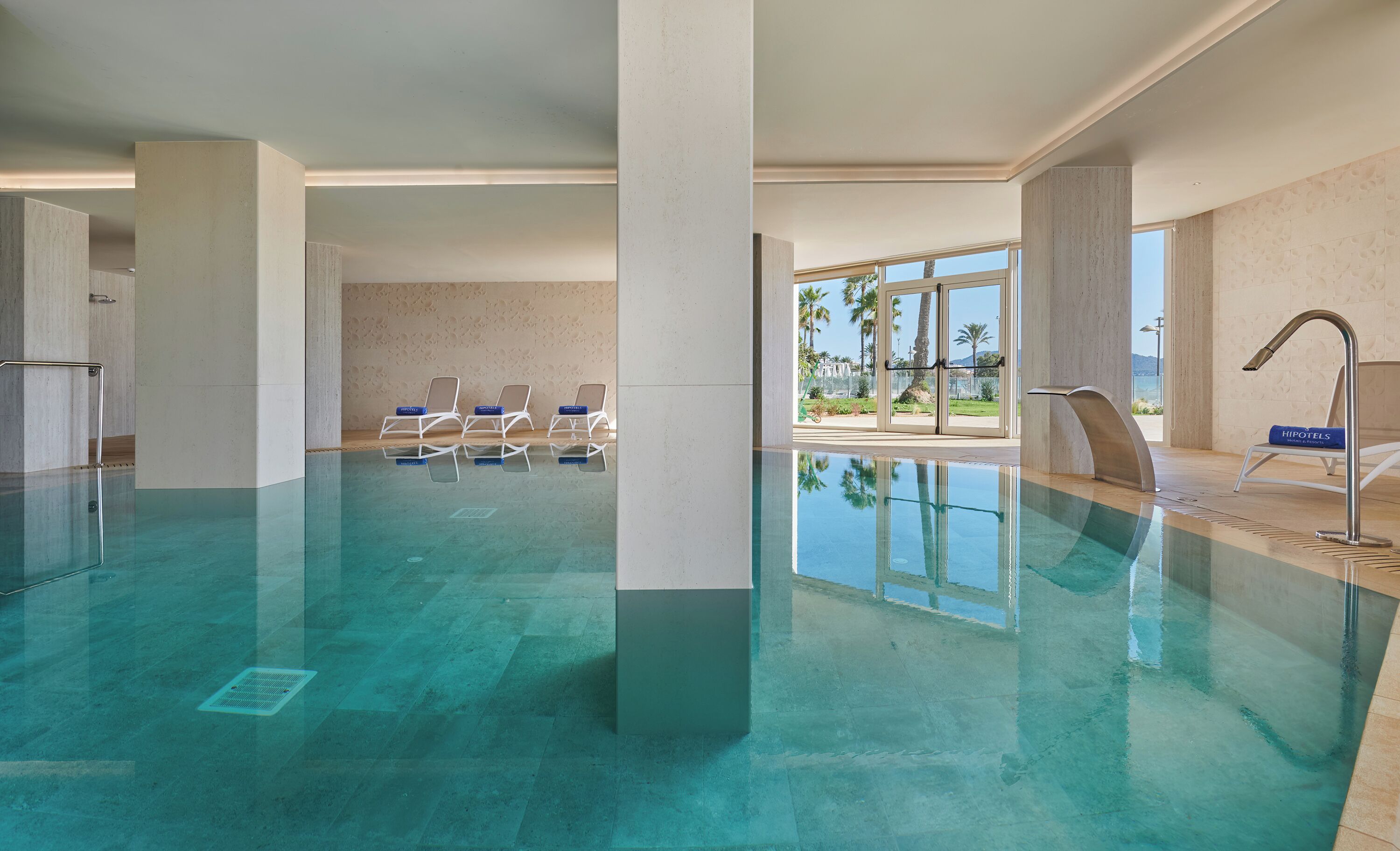 A swimming pool with clear water, surrounded by white columns and a view of palm trees and mountains.