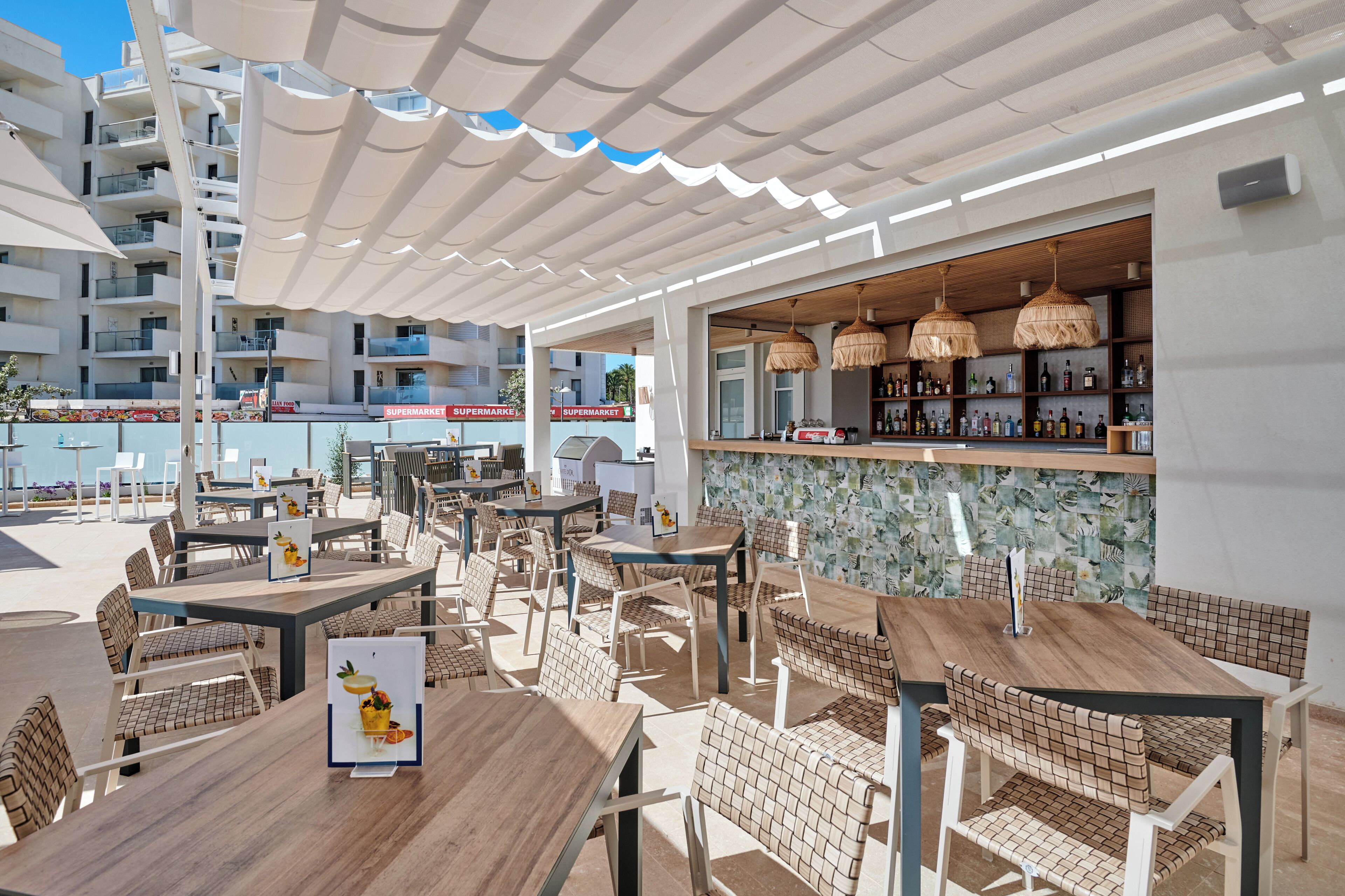 An outdoor seating area with tables and chairs, a bar with shelves of bottles, and a view of a supermarket in the background.