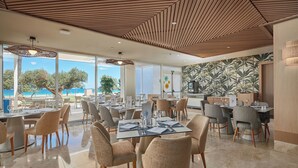 Un restaurante moderno con vistas al mar, que cuenta con techo de madera, ventanas amplias y un diseño de papel tapiz tropical.