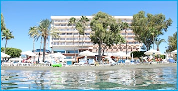 Hotel à beira-mar com várias varandas, palmeiras e guarda-sóis de praia.
