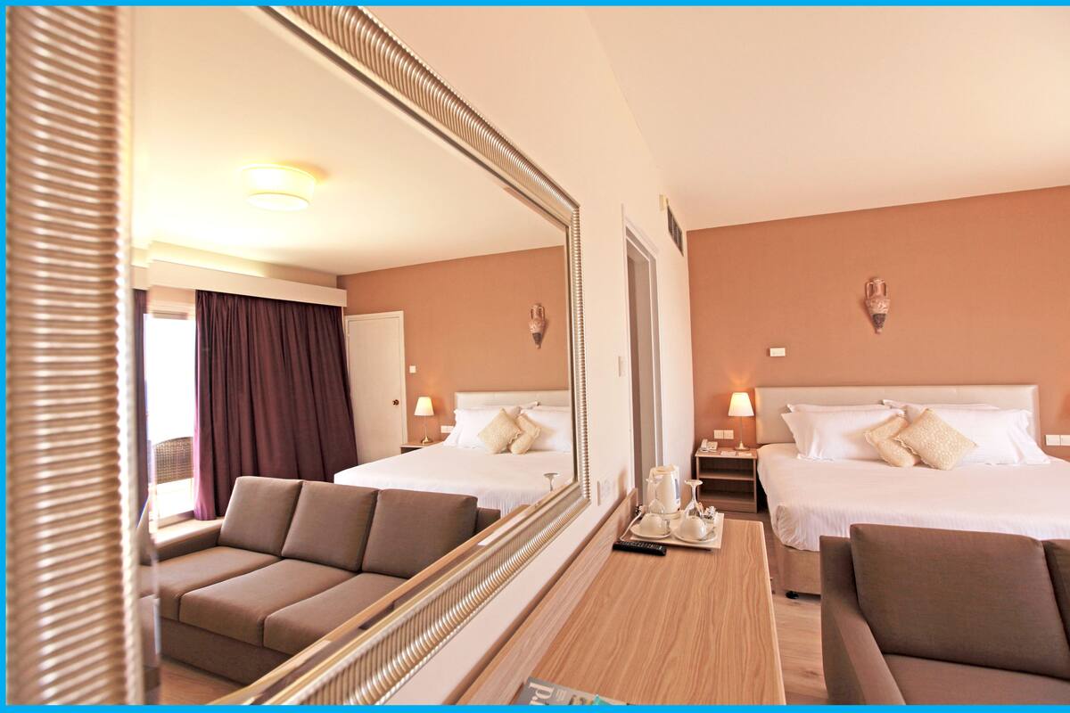 Ein Hotelzimmer mit zwei Betten, einem Sofa, einem Schreibtisch mit Lampe und einem großen Spiegel.