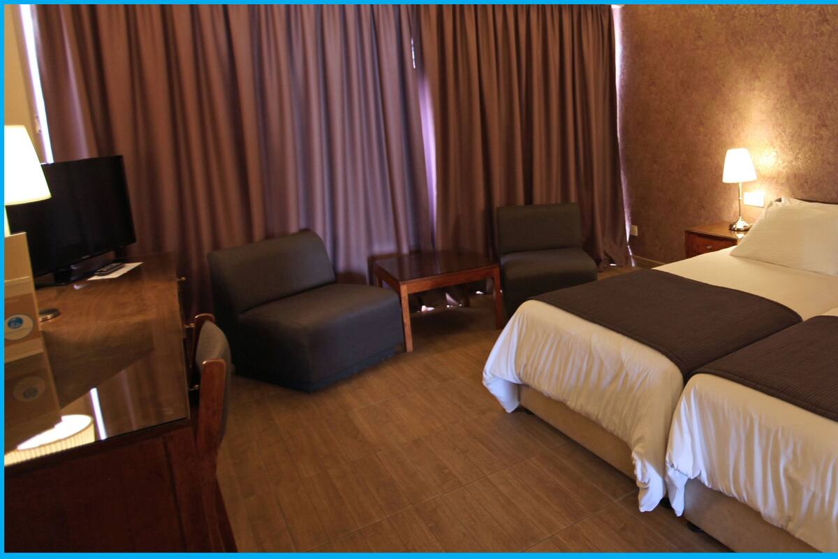 Ein Hotelzimmer mit einem großen Bett, zwei Nachttischen mit Lampen, einer kleinen Couch, einem Fernseher und Vorhängen.