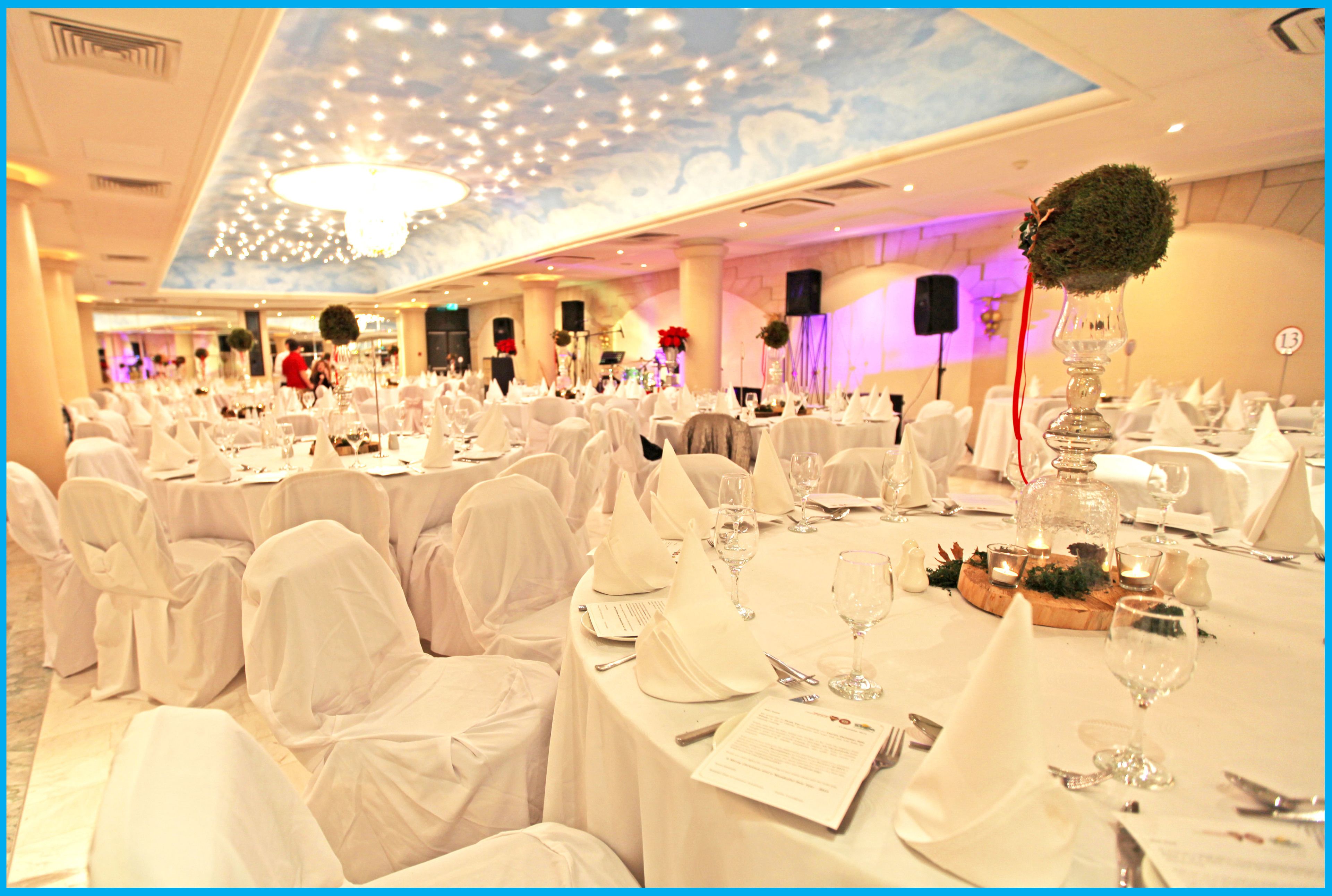 A banquet hall with round tables covered in white tablecloths, chairs with white covers, and a chandelier.