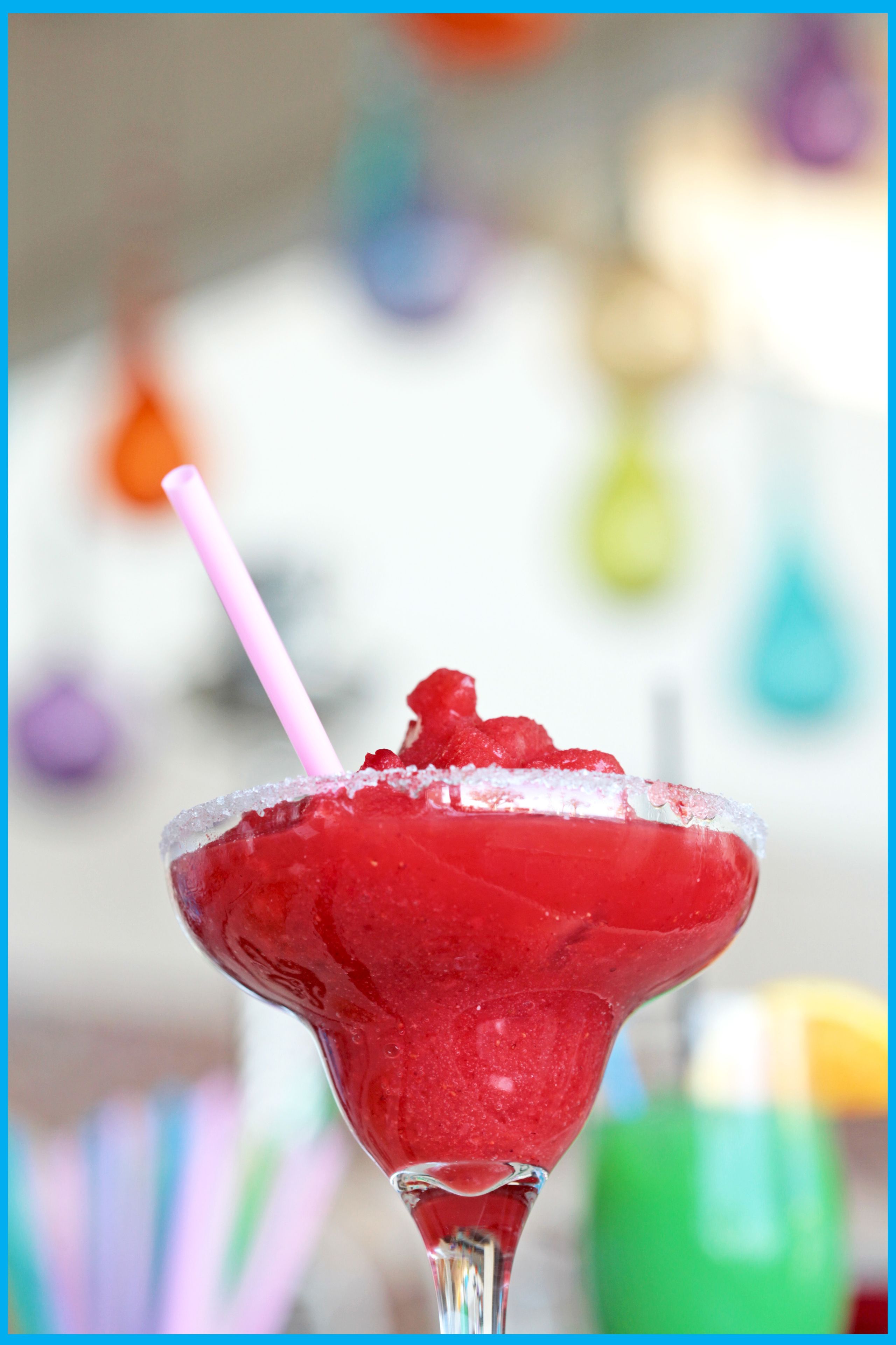 Primer plano de un cóctel rojo con una pajita rosa en una copa de martini, con un fondo borroso de decoraciones coloridas.