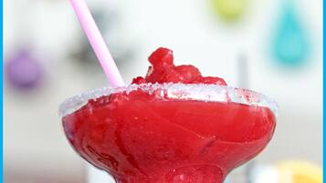 Primer plano de un cóctel rojo con una pajita rosa en una copa de martini, con un fondo borroso de decoraciones coloridas.