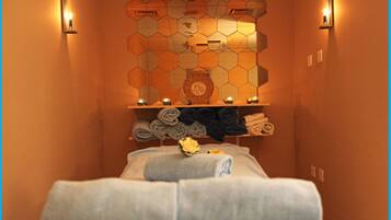 Relax in a serene spa room with a central bed, surrounded by a geometric patterned wall and candles, and convenient storage for your essentials.