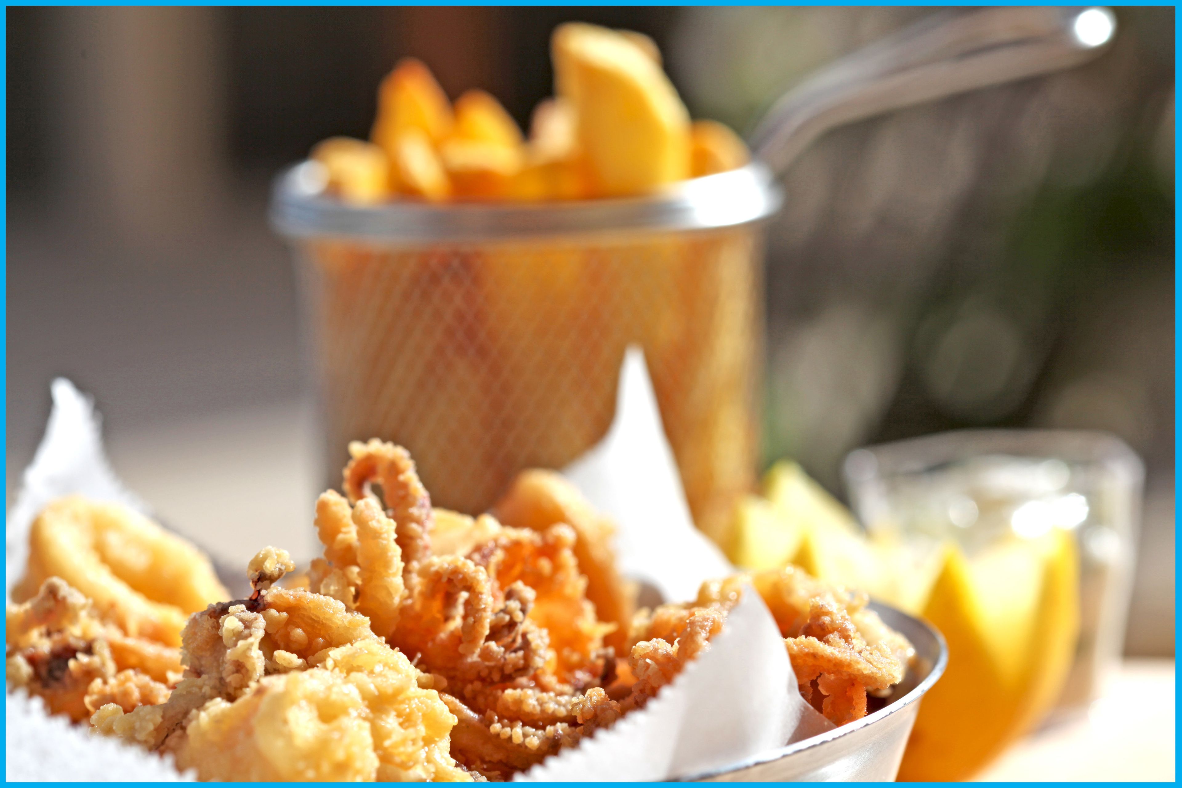 A basket of fried food, likely calamari, with lemon wedges and a glass of a clear beverage, possibly water or juice, on a table.