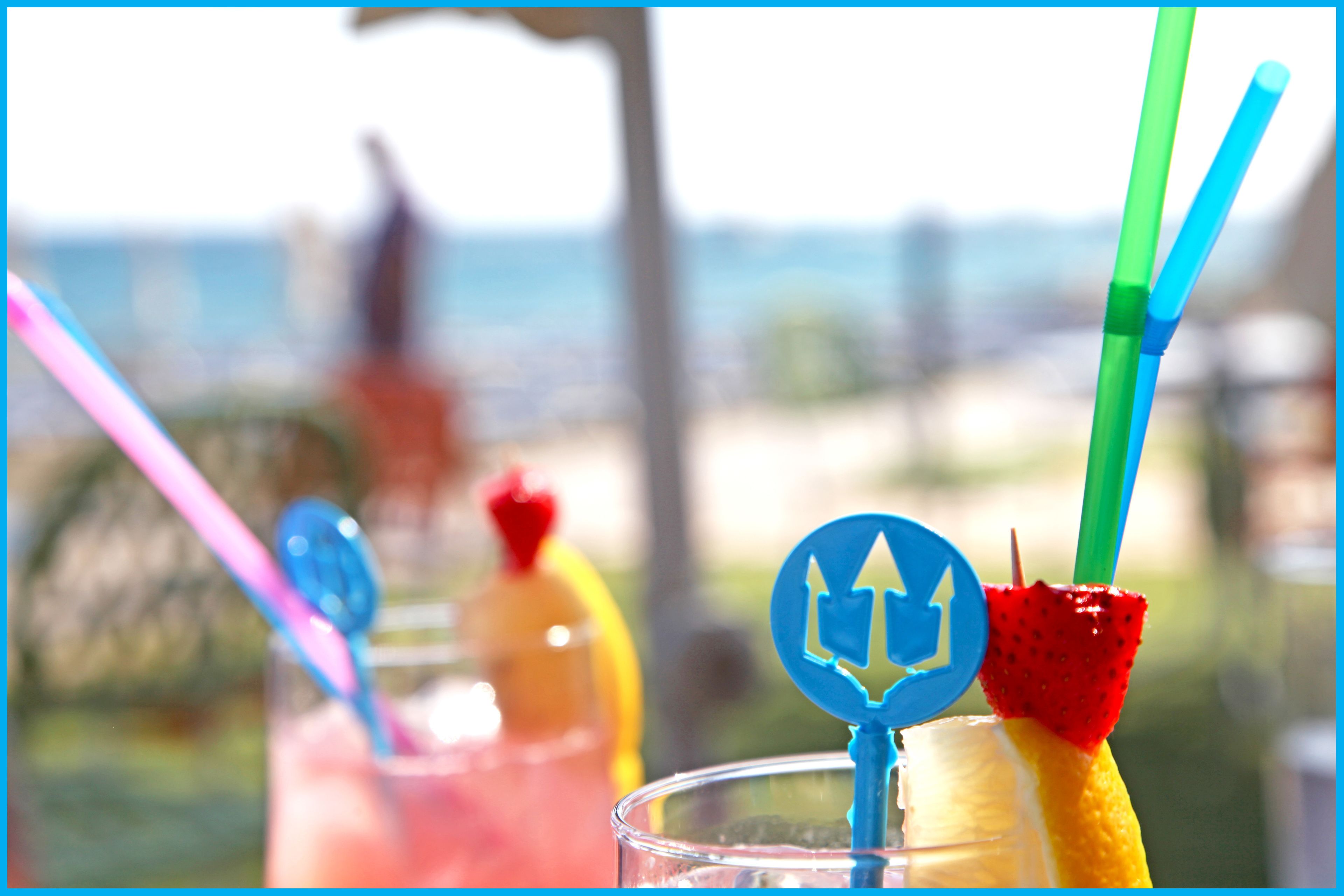 A glass with a straw and a fruit garnish, with a blurred background suggesting an outdoor setting.