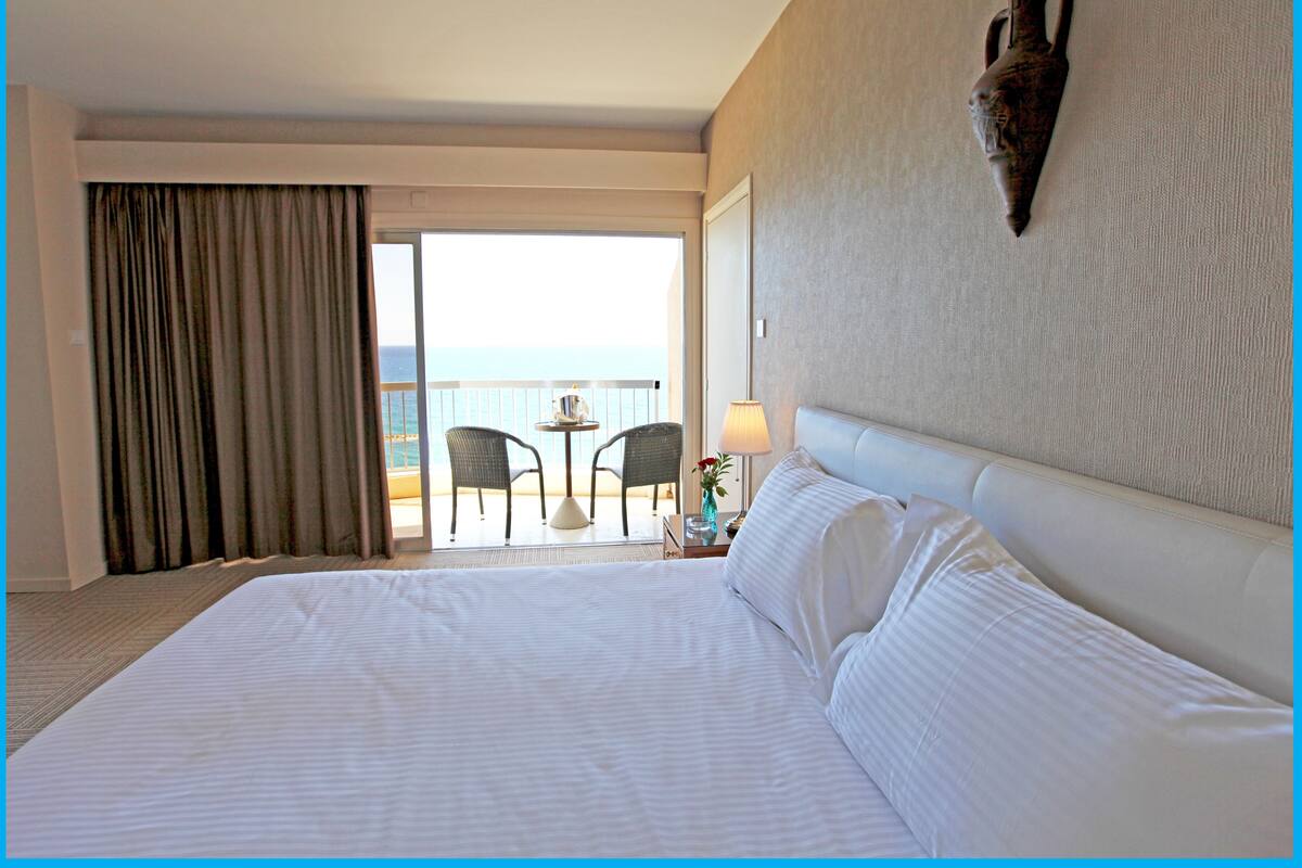Ein Hotelzimmer mit einem großen Bett, einem Balkon mit Sitzgelegenheiten und Meerblick.