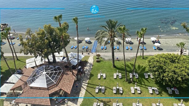 Vista aérea de un complejo turístico frente a la playa, con palmeras, tumbonas y un pabellón.