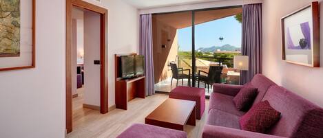 Habitación de hotel con balcón, sofá, televisor y zona de comedor con mesa y sillas.