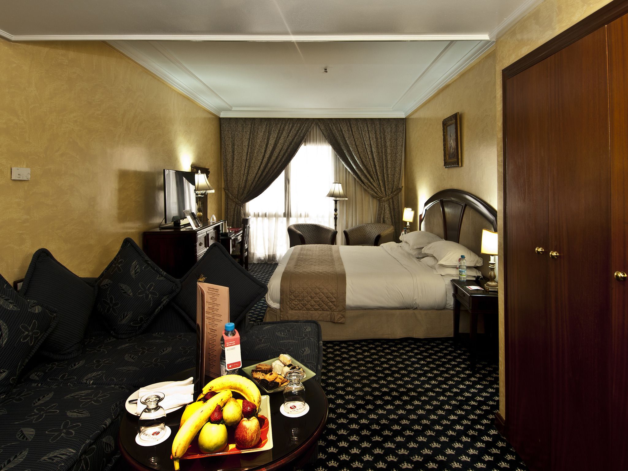 Habitación de hotel con cama, escritorio, silla, mesa de comedor con frutas y una botella, y un armario.