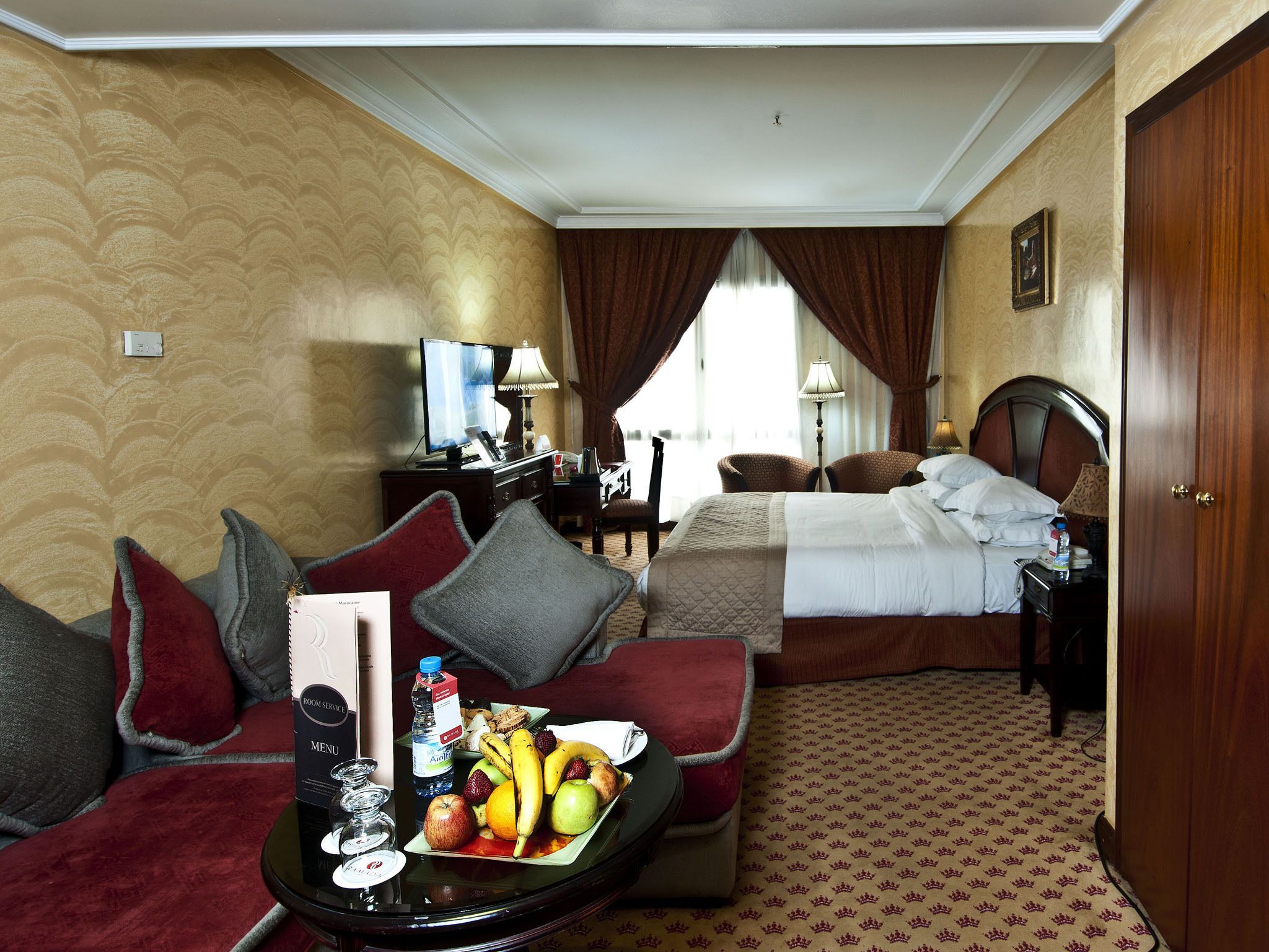 A hotel room with a bed, a sofa, a desk with a computer, and a dining table with fruit and a menu.