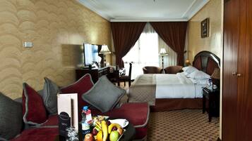 A hotel room with a bed, a sofa, a desk with a computer, and a dining table with fruit and a menu.
