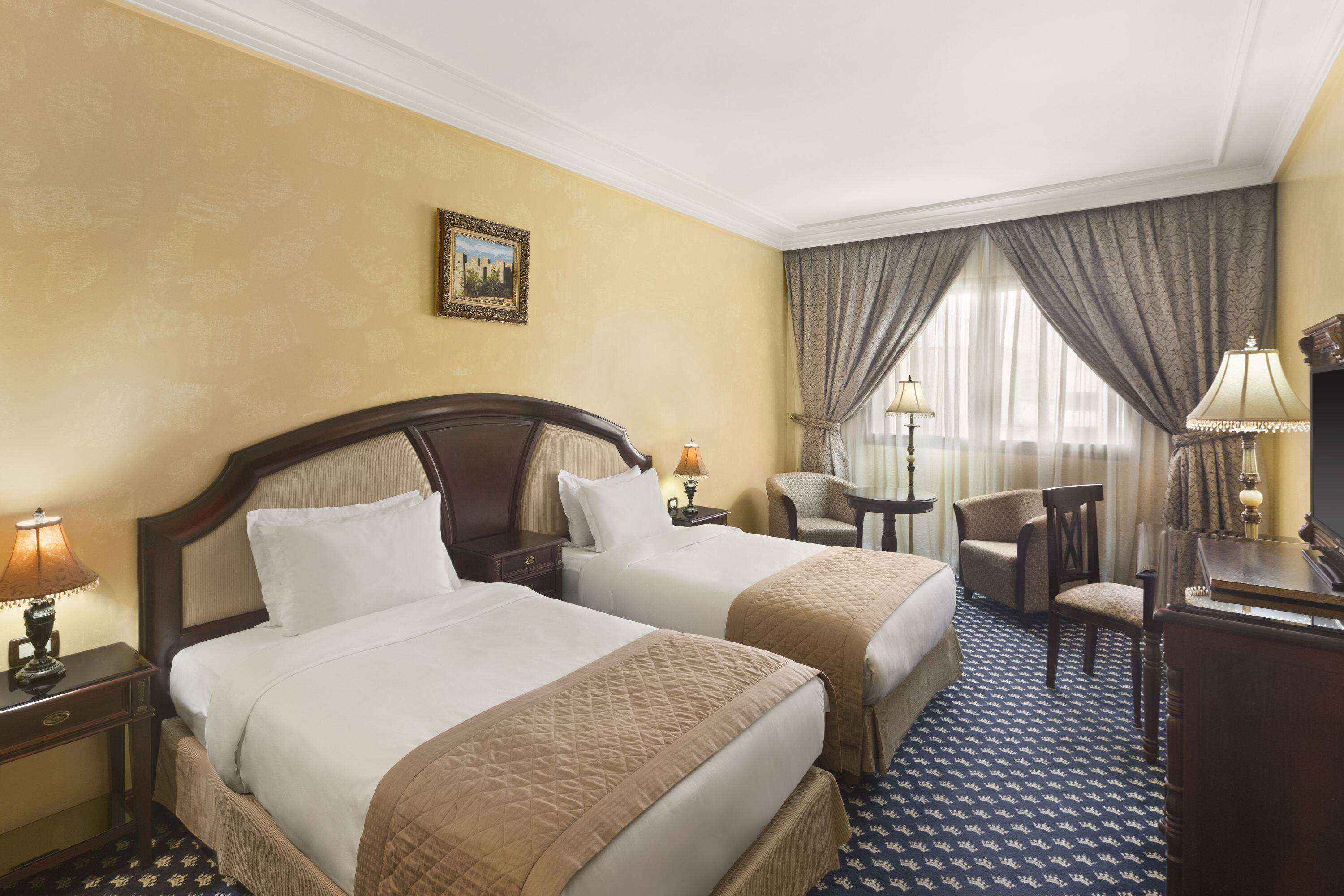 A hotel room with two beds, a desk, a chair, and a lamp.