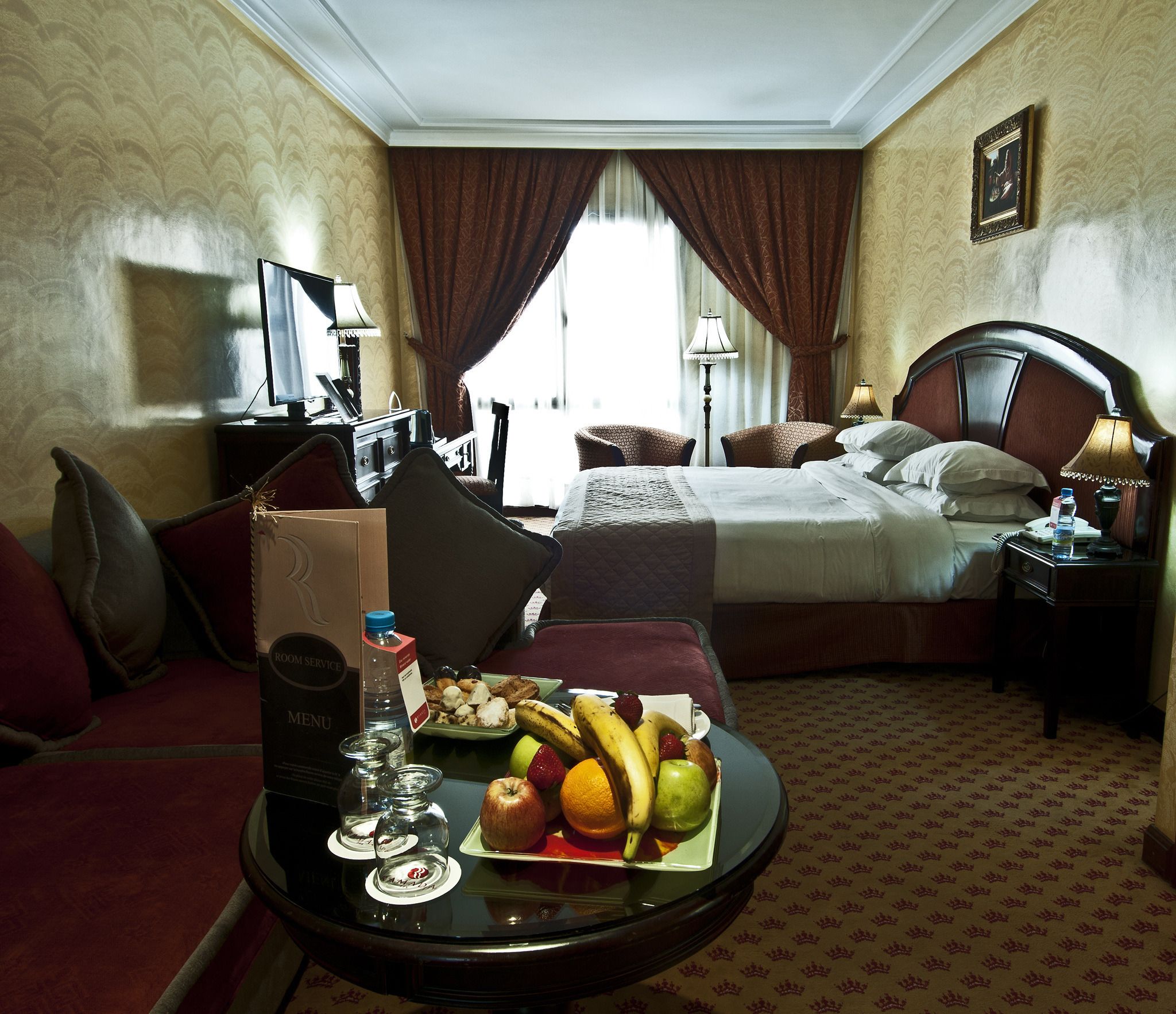 A hotel room with a bed, bedside table, and a dining table with fruit and a water bottle.