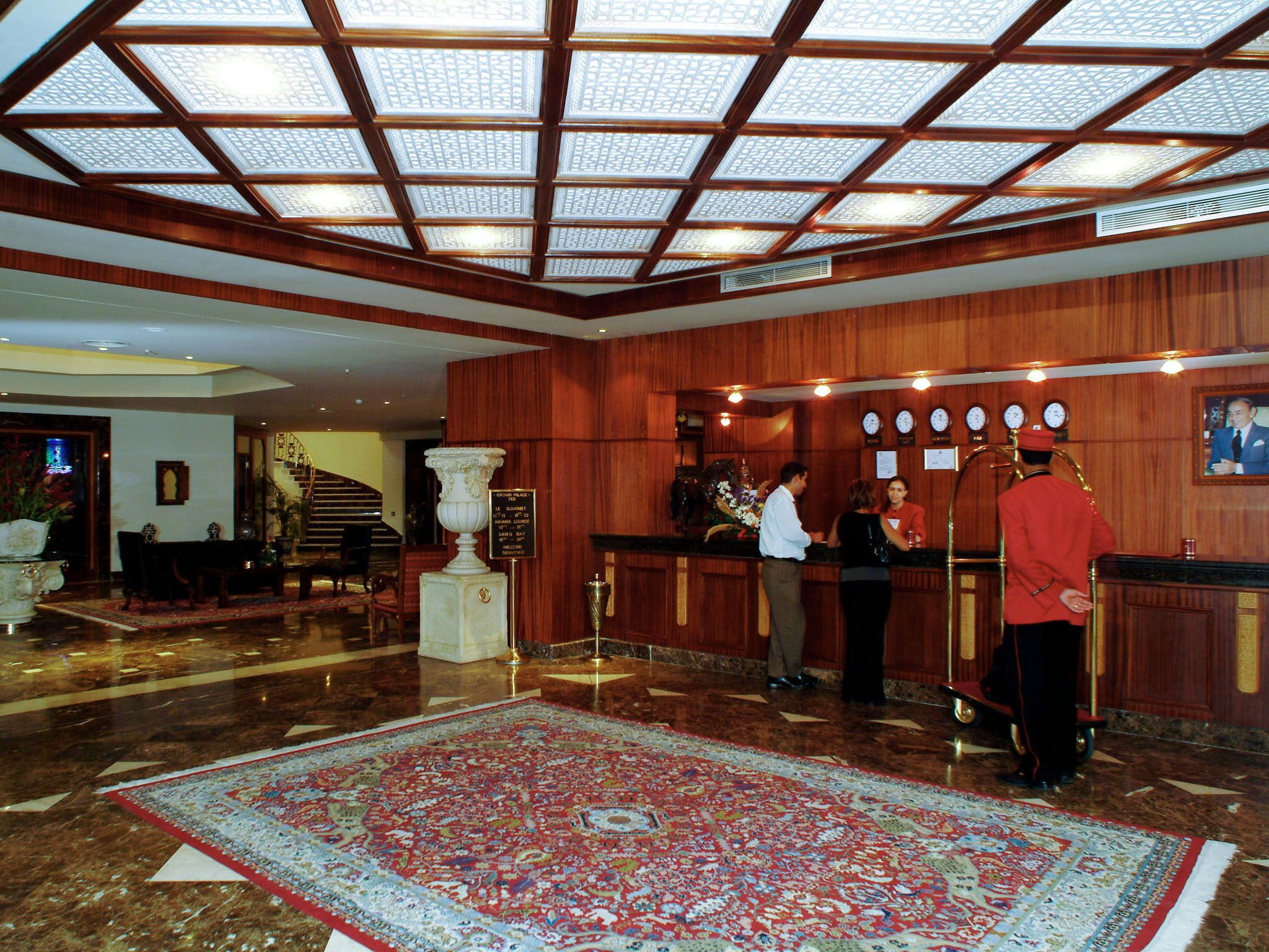 A hotel lobby with a large wooden reception desk, a grand staircase, and a patterned carpet.