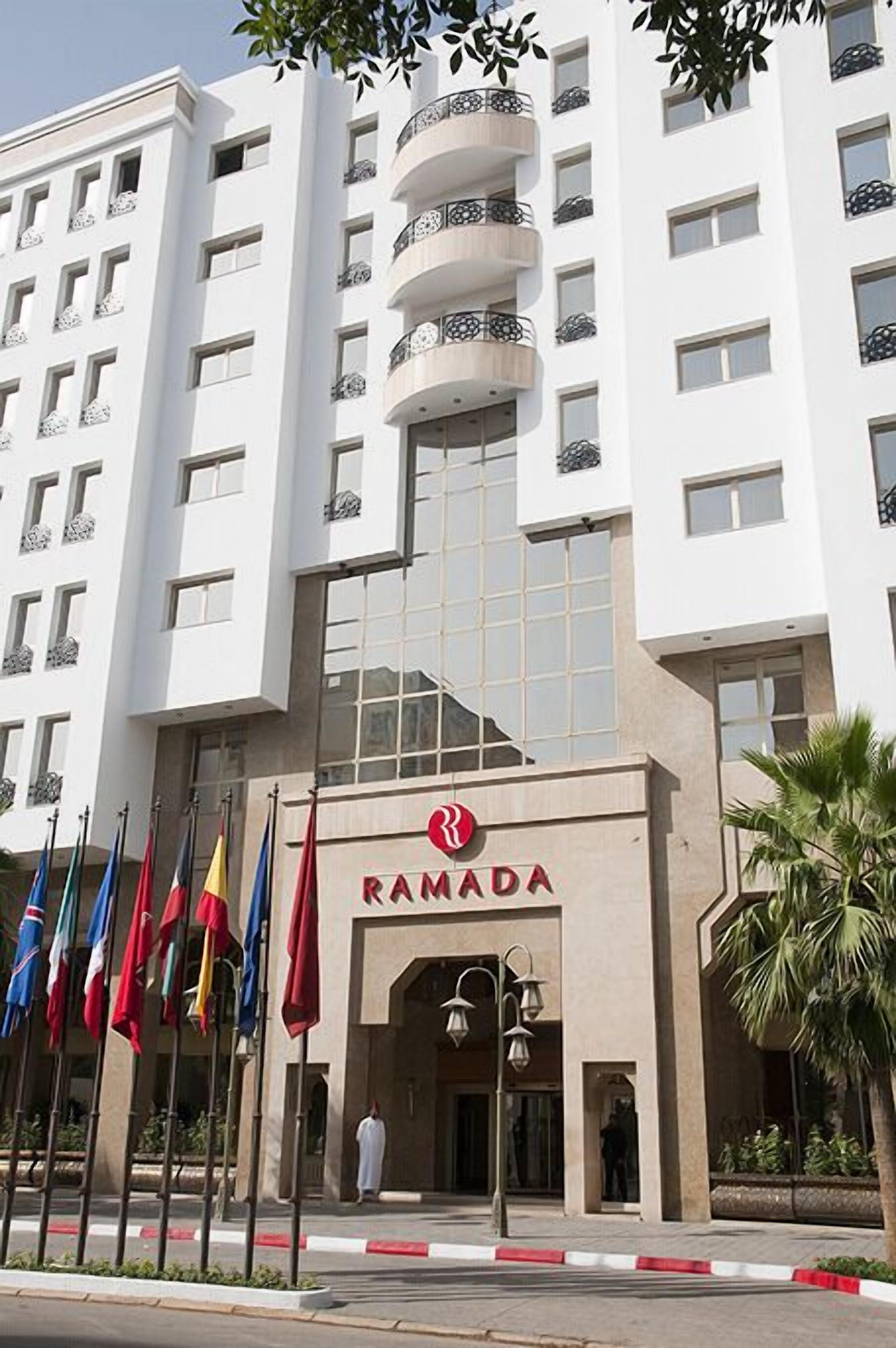 Ein modernes Gebäude mit einem gut sichtbaren „RAMADA“-Schild, mehreren Balkonen und Wimpeln davor.
