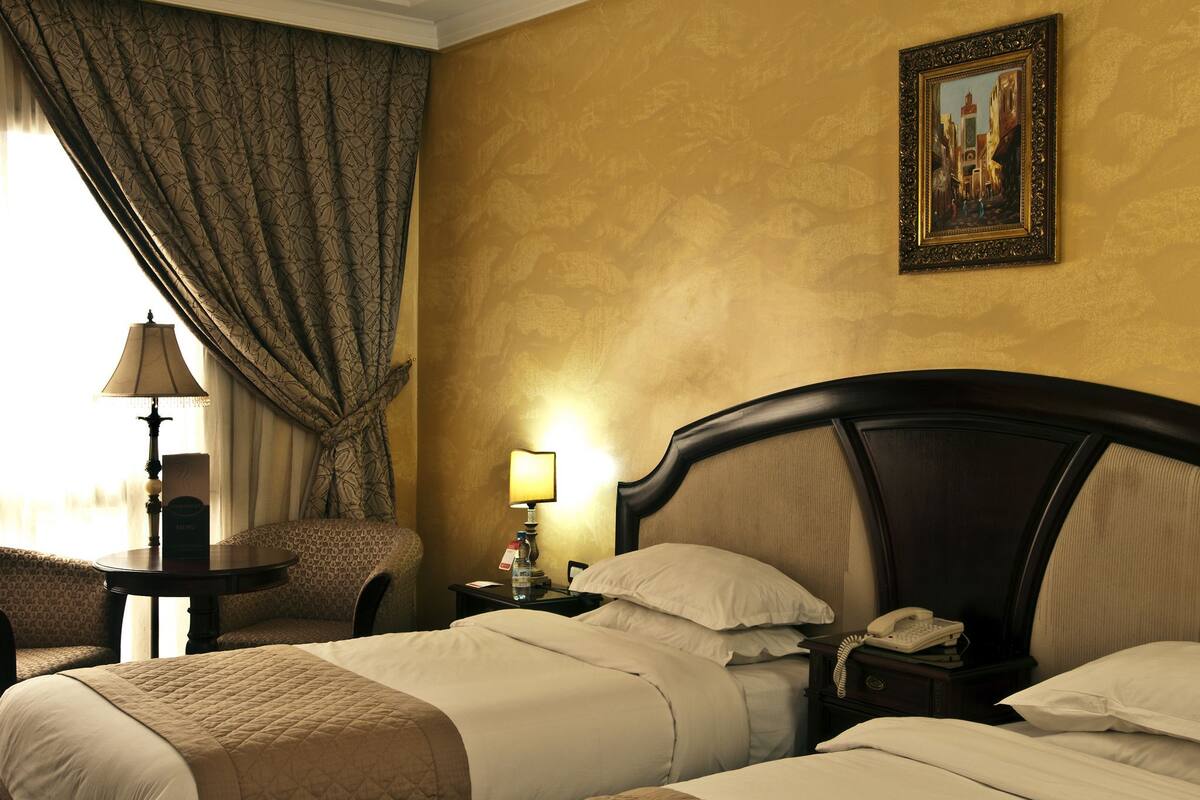 Quarto de hotel com duas camas, uma mesa de cabeceira com abajur, uma cadeira e um quadro na parede.