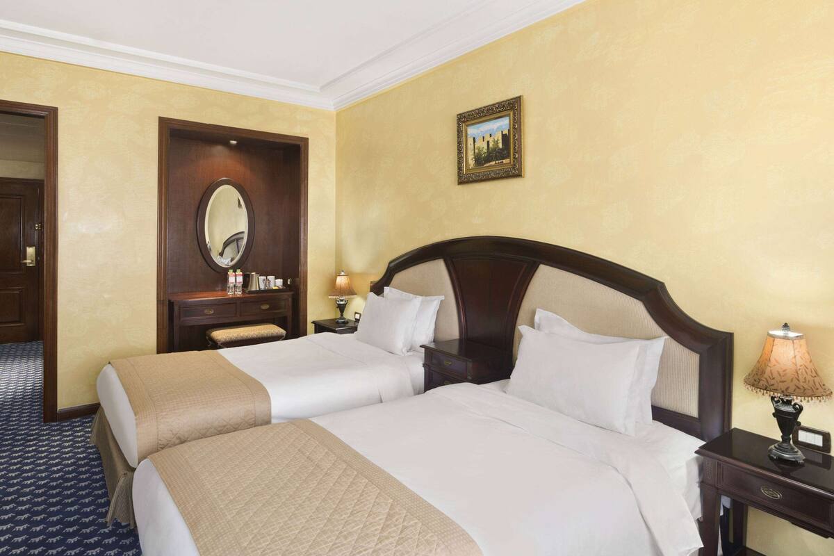 Quarto de hotel com duas camas, cabeceira de madeira, mesa de cabeceira com abajur, espelho e quadro na parede.
