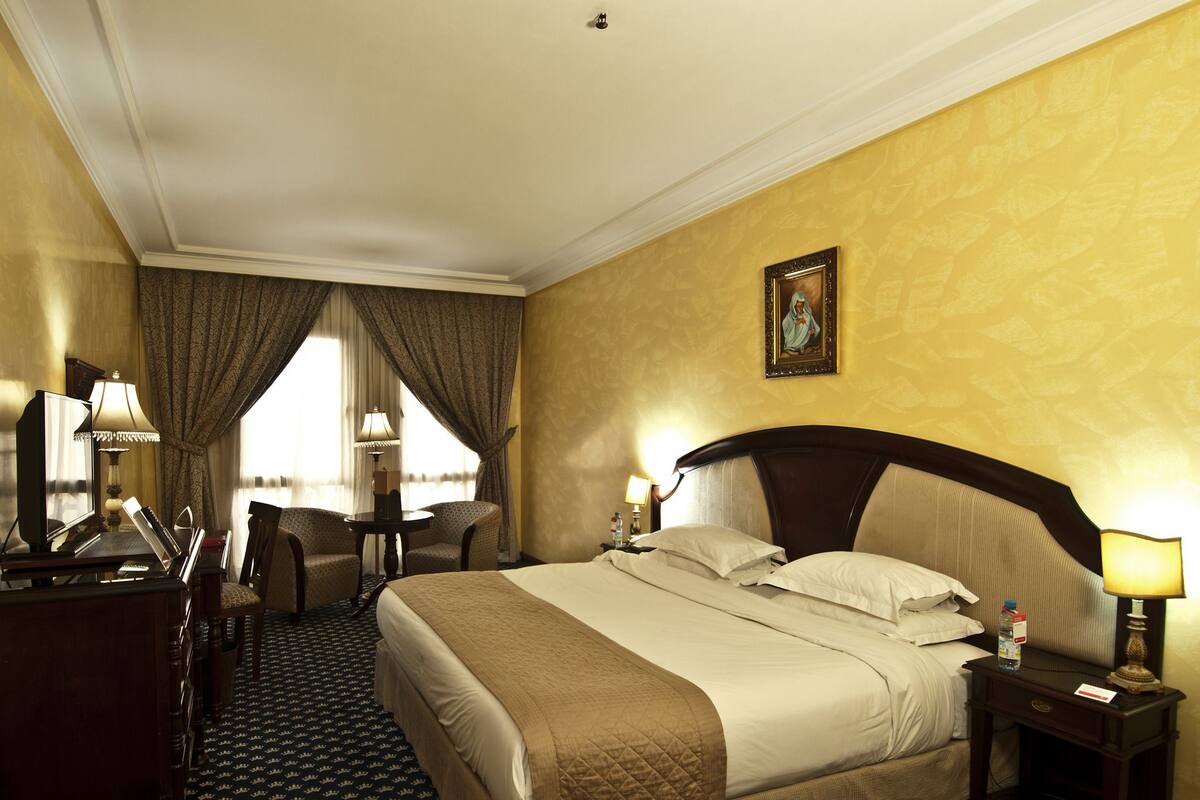 Quarto de hotel com uma cama grande, uma escrivaninha com luminária, uma cadeira e um quadro na parede.